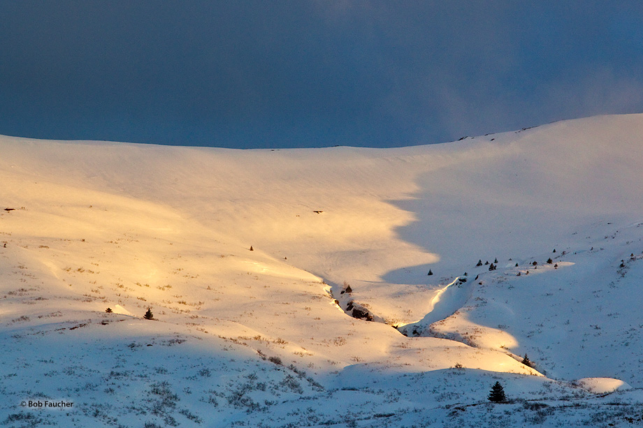 A break in the ever-present cloud cover reveals cobalt blue skies with wisps of blowing snow over a high river valley whose exposed...