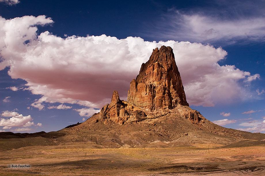 A huge cumulus cloud passing near Agathla Peak has its undersurface colored by the reflection off the desert sands below