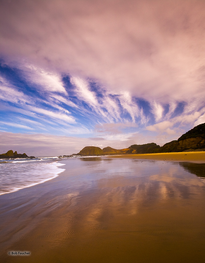 Streaking clouds overhead and their reflections in the wet sands of the beach appear to converge on the headlands