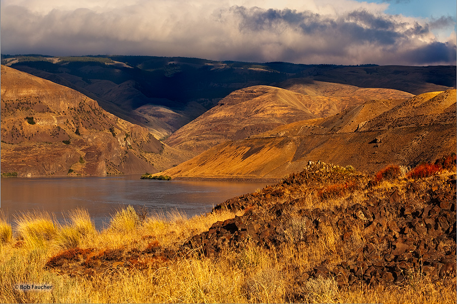 The Columbia River meanders through the upper Columbia Gorge, flanked by rocky hills bathed in dawn's first light while storm...