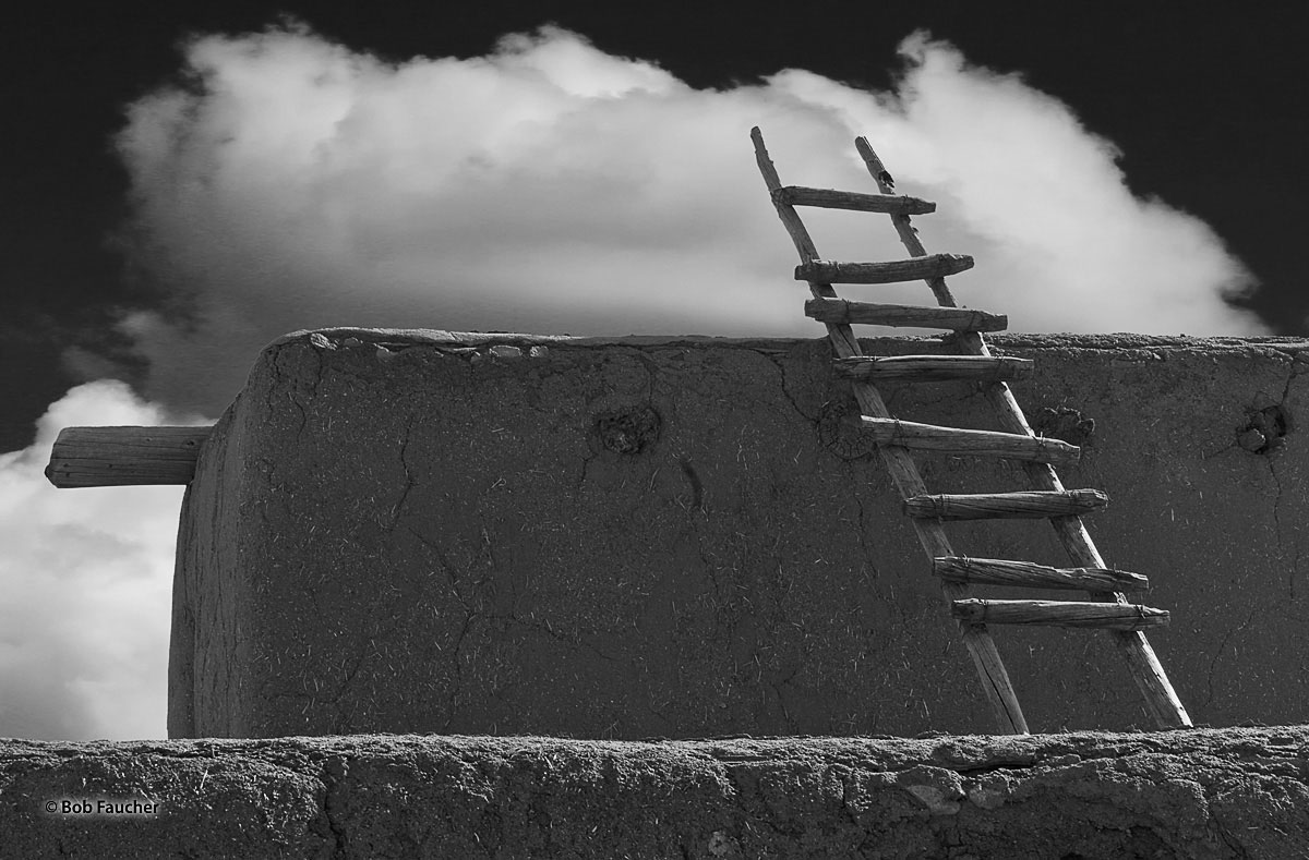 A wooden ladder reaches up from the second floor roof to the third story roof of one of the historical adobe buildings forming...