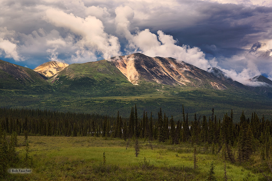 Evening sun breaks under early Summer storm clouds hanging over the strikingly striped hills along the Old Denali Highway in...