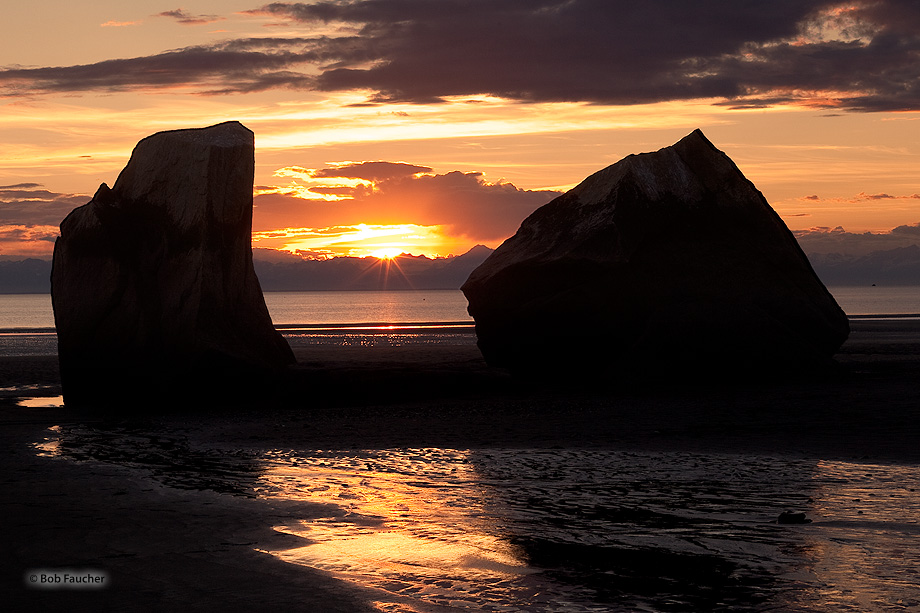 Sunset and low tide are coincident on this late summer evening. The glow of the sun rays passing between the boulders reflects...