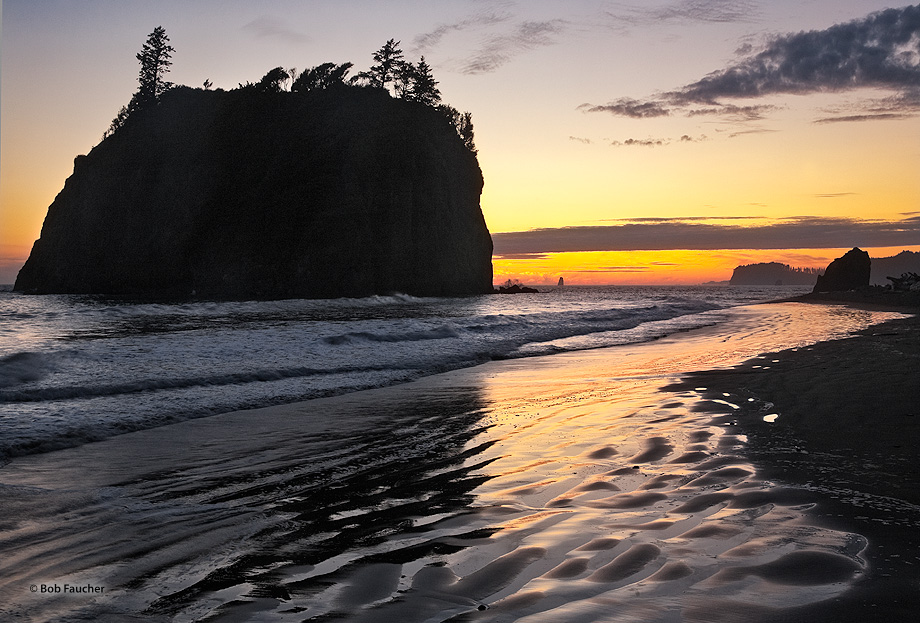 Setting sun silhouettes the sea stack casting shadows and forming reflections on the wet rippled sand at Ruby Beach