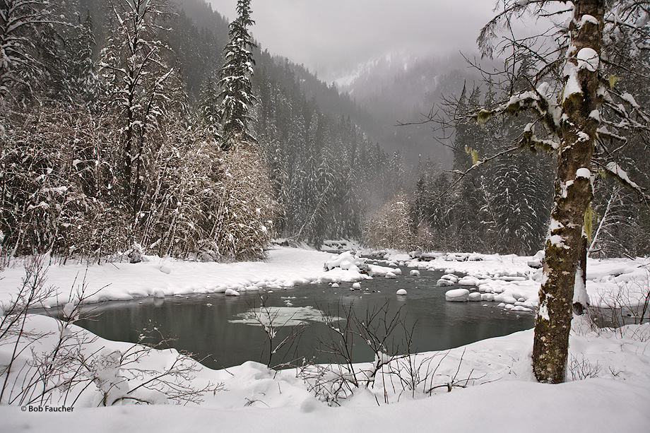 Partially frozen river with snow covered banks and hummocks meanders into the cloudy, fog-shrouded valley