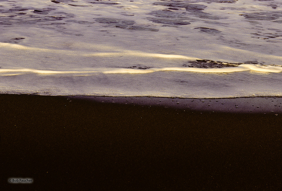 Sea foam highlighted by early morning sun and contrasting with the black sand of Waimea Beach